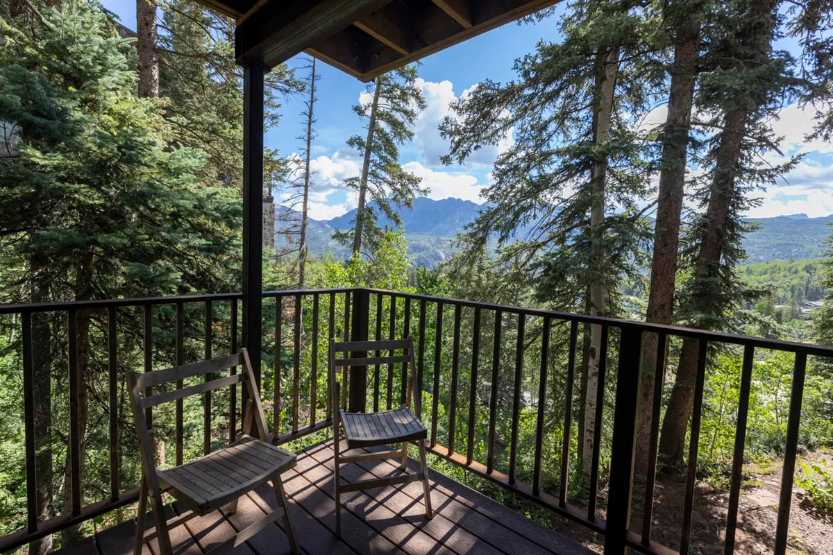 A peaceful seating area with wooden chairs faces a sweeping mountain vista framed by trees, ideal for relaxation after a day on the slopes. The sunset light bathes the scene, evoking tranquility and inviting views from the property’s balcony or patio.