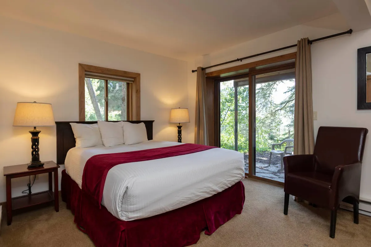 A spacious bedroom with a king-size bed is complemented by a large sliding glass door that opens to a balcony. From the bed and armchair, guests can enjoy serene sunrise views over the forest, inviting them to start each day with a connection to nature.