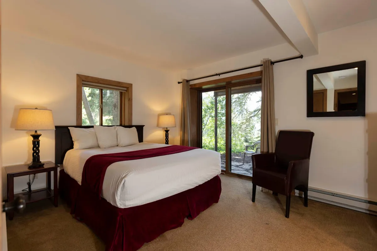 A serene bedroom featuring a king-sized bed with a burgundy throw, two bedside lamps casting a warm glow, a sliding glass door that opens to an outdoor area, and a leather armchair. The room is inviting, restful, and offers tranquil views that connect the indoors to nature.