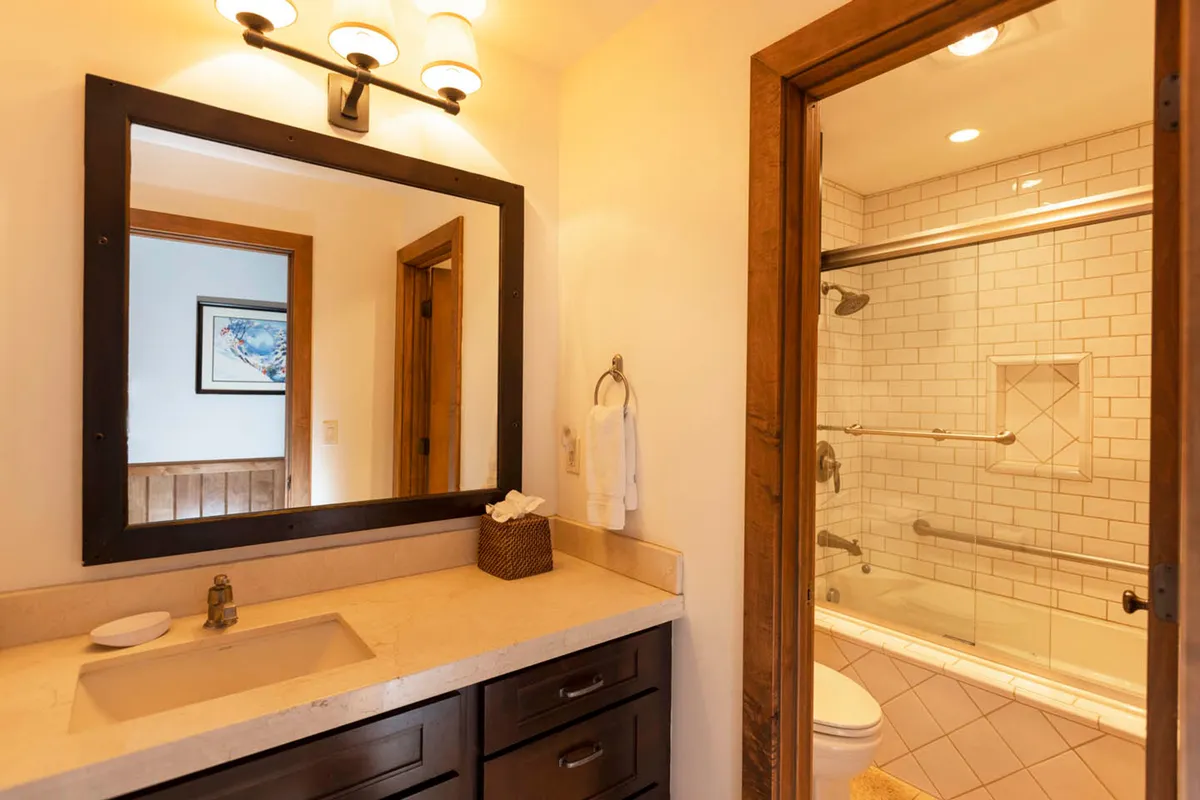 A warm, inviting bathroom with a large framed mirror above a sink and countertop, wooden cabinetry, and a glimpse of the bathtub and toilet area through the doorway. The space is well-lit and features natural wood finishes that add a touch of rustic charm.