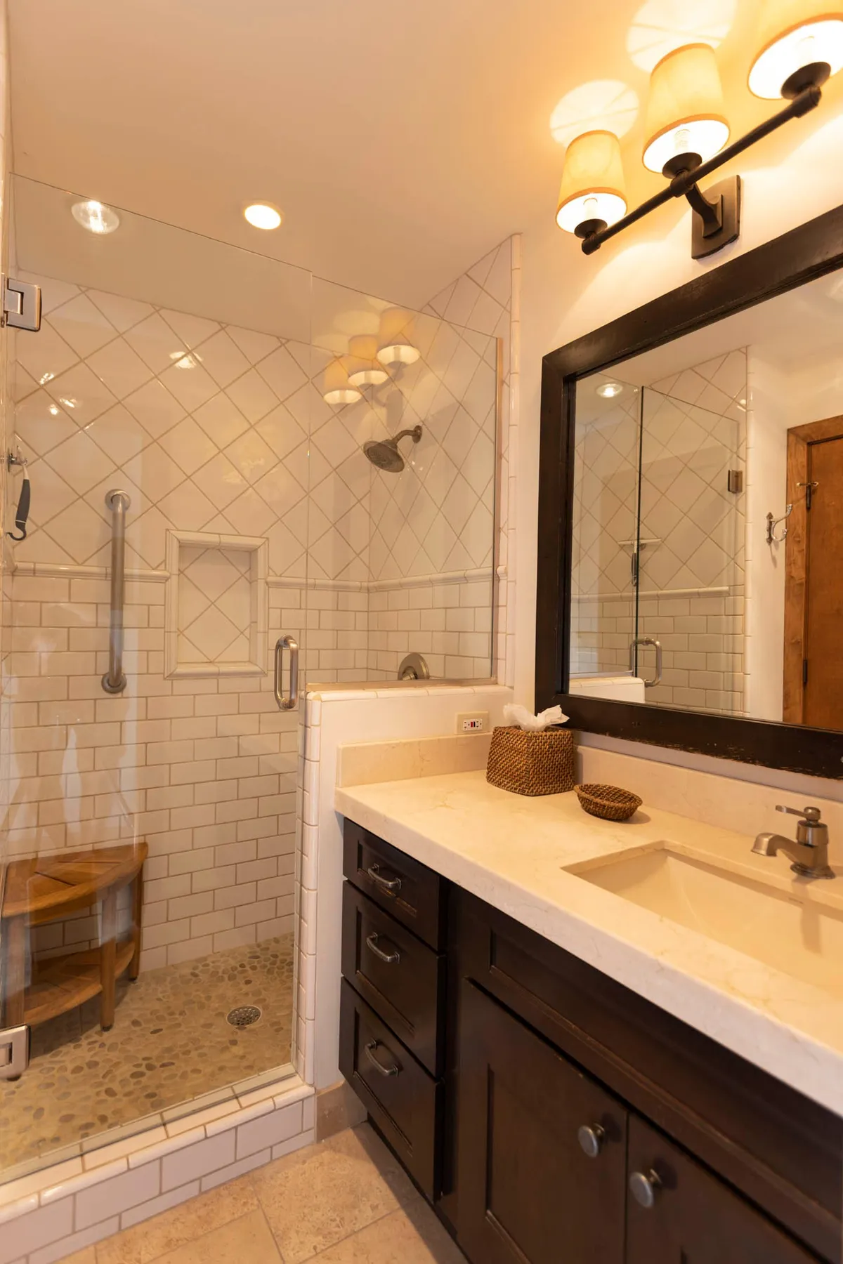 A bright, modern bathroom featuring a glass-enclosed shower with a pebble floor and wooden bench, a dark wood vanity with a beige countertop, a large mirror, and soft wall-mounted lighting. The room exudes a spa-like, relaxing ambiance.