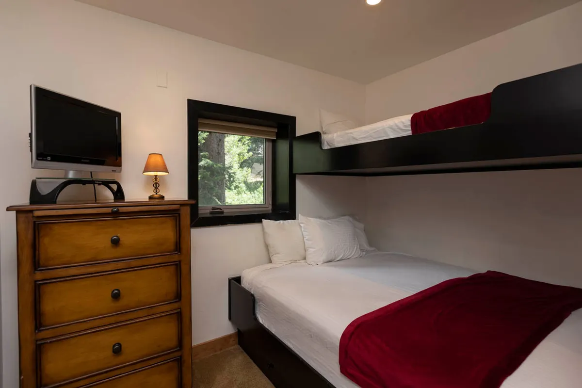 A cozy bunk room featuring bunk beds with red blankets, a small window with forest views, a wooden dresser with a lamp, and a wall-mounted TV. The space feels snug and intimate—ideal for kids or friends seeking a comfortable, fun sleepover environment.