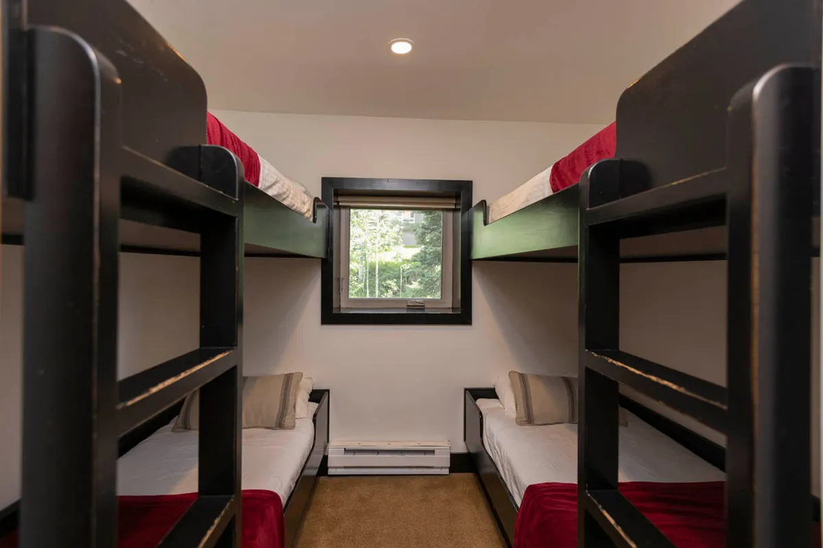 A practical but inviting bunk room with black wooden bunk beds, small window offering a forest view, red bedding accents, and neutral walls and flooring. The space is clearly designed for shared sleeping, perfect for kids or groups.