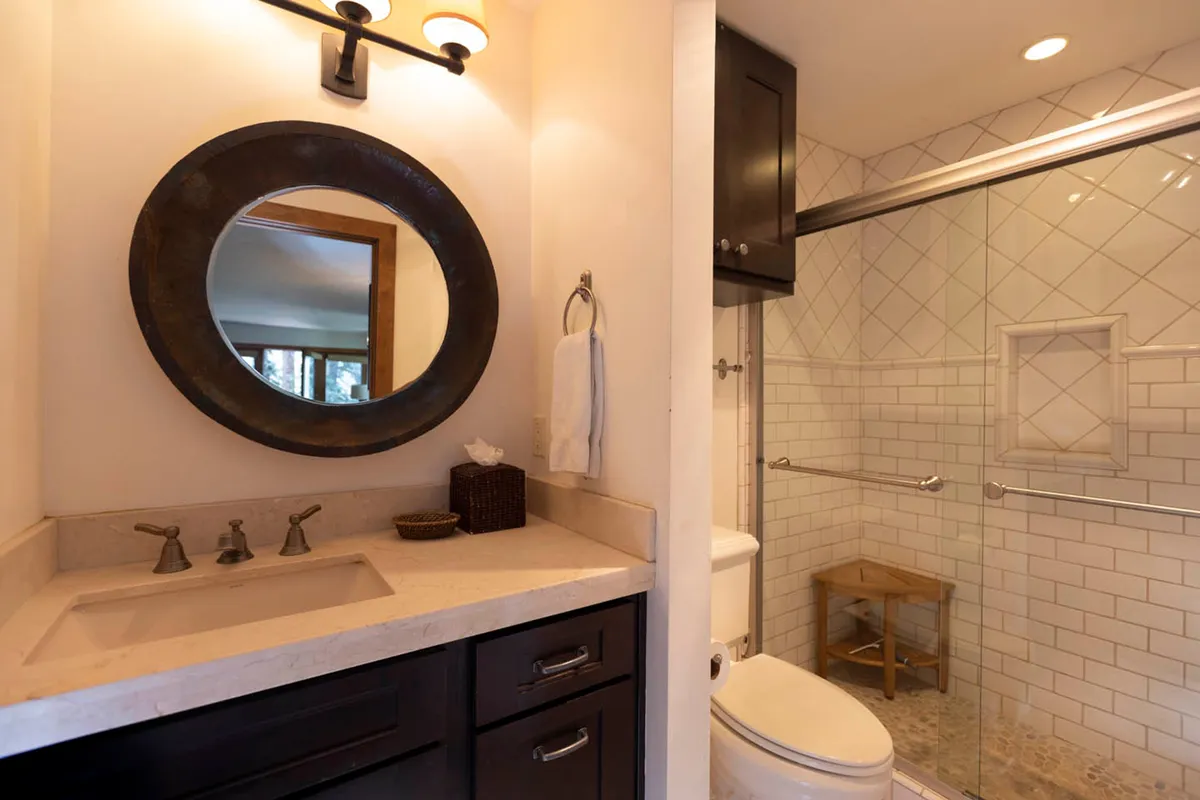A sleek bathroom showcasing a round decorative mirror, a vanity with a beige countertop and dark wood drawers, a glass-enclosed shower with tiled walls, and a toilet. The space feels calm, organized, and practical, with subtle decorative touches.