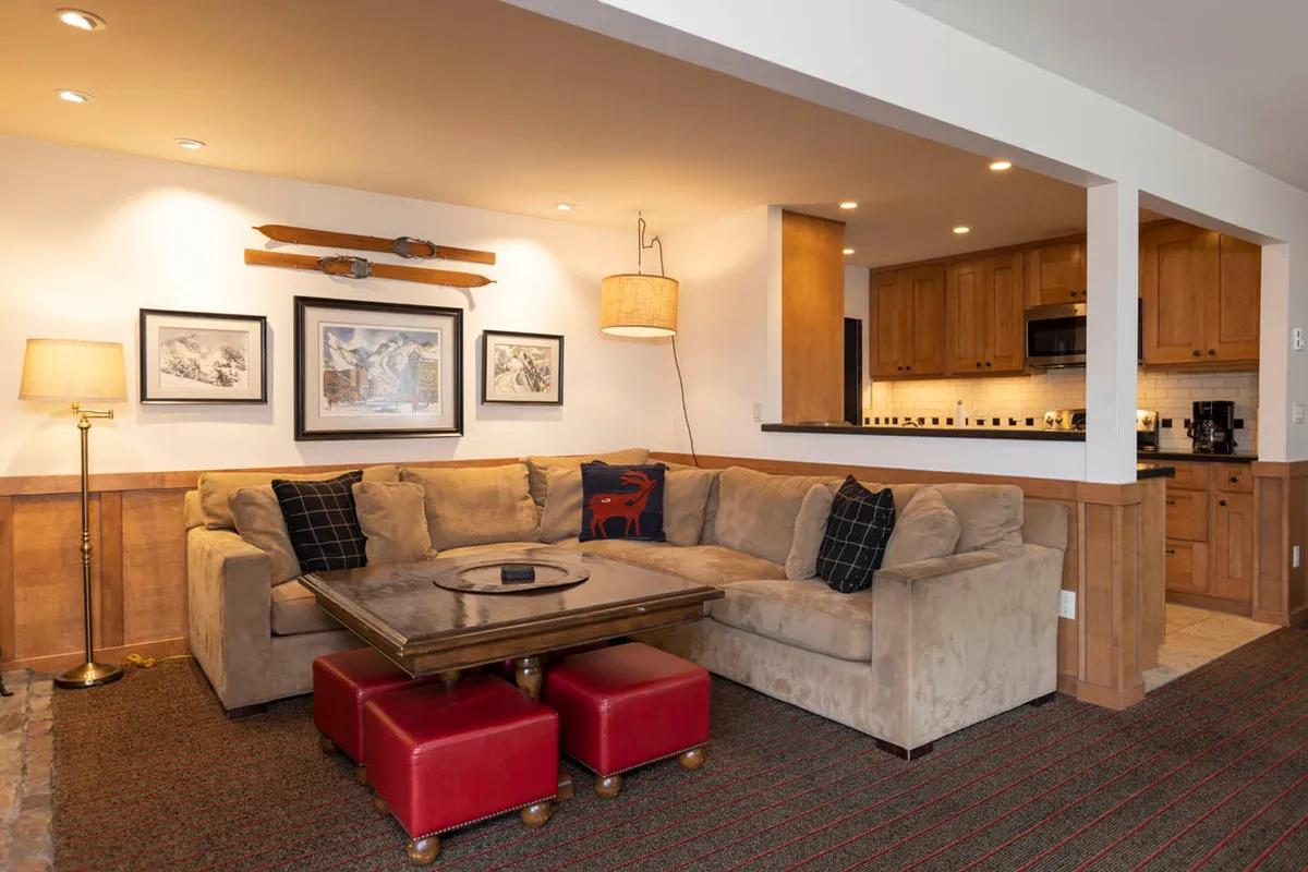 A spacious open-concept living area with a large beige sectional sofa, wooden coffee table, and red ottomans. The focal wall features vintage skis and mountain-themed artwork, and the space feels cozy and inviting for group relaxation. The living area connects visually to the kitchen and dining zone, suggesting a unified hub for après-ski gatherings.