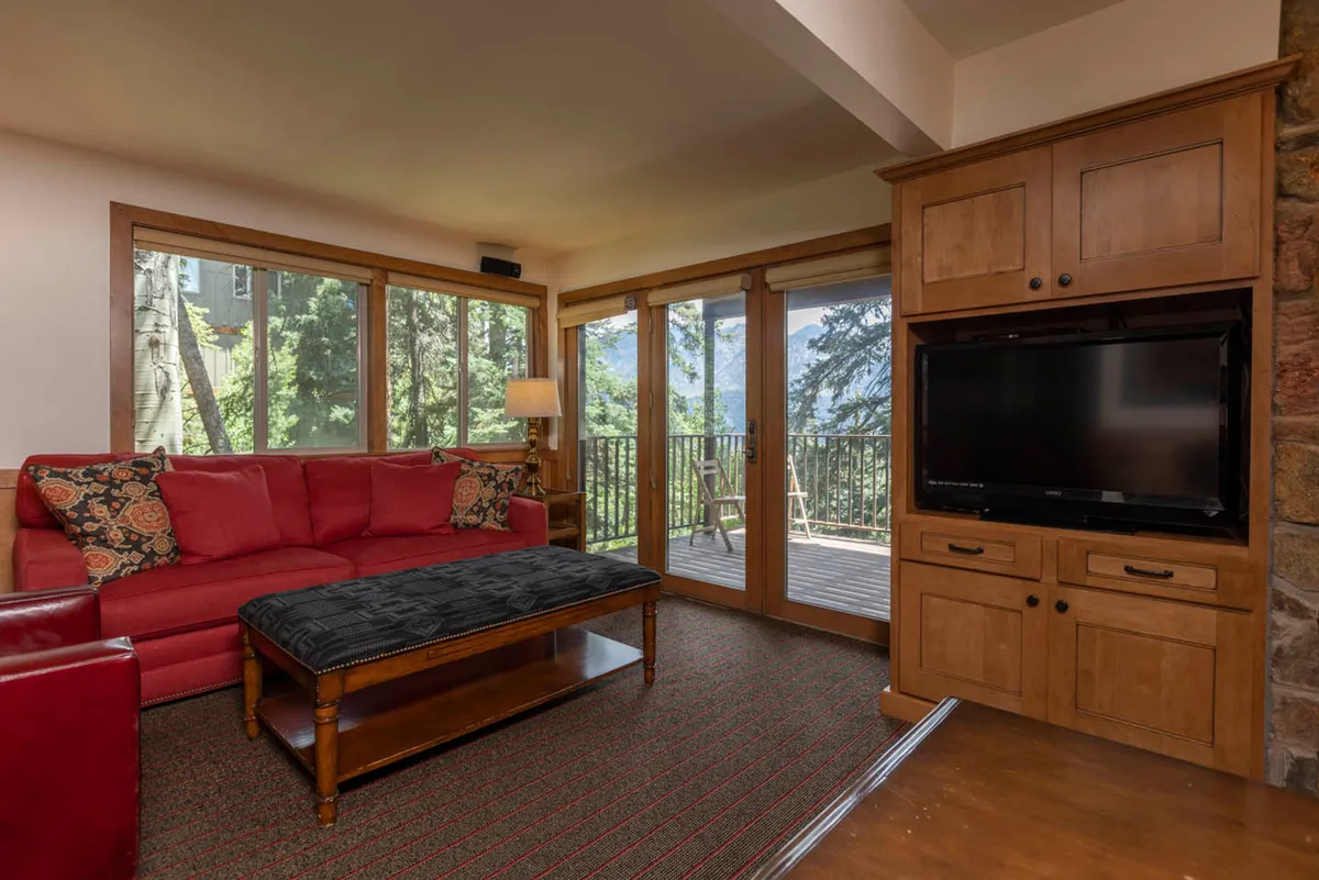 A cozy living area with a red sofa, patterned pillows, and a large flat-screen TV sits near a balcony with expansive views of forests and mountains. The inviting indoor space is seamlessly connected to the outdoors, offering opportunities to relax after a day of adventure while soaking in sunset vistas.