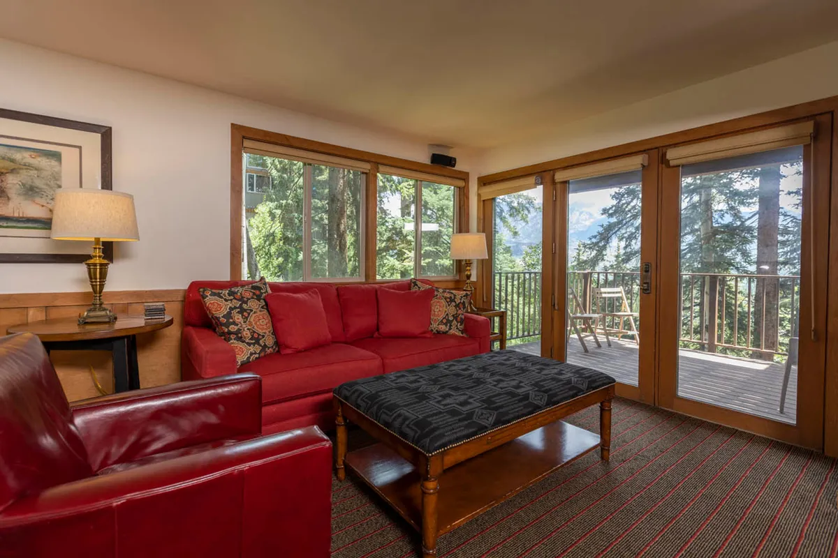 A cozy living area featuring a red sofa adorned with patterned pillows, a black patterned ottoman serving as a coffee table, and large glass doors that open to a balcony with wood-framed windows offering scenic forest views. The soft lighting and comfortable seating invite group gatherings and relaxation, with easy access to the outdoors for fresh air and mountain scenery.