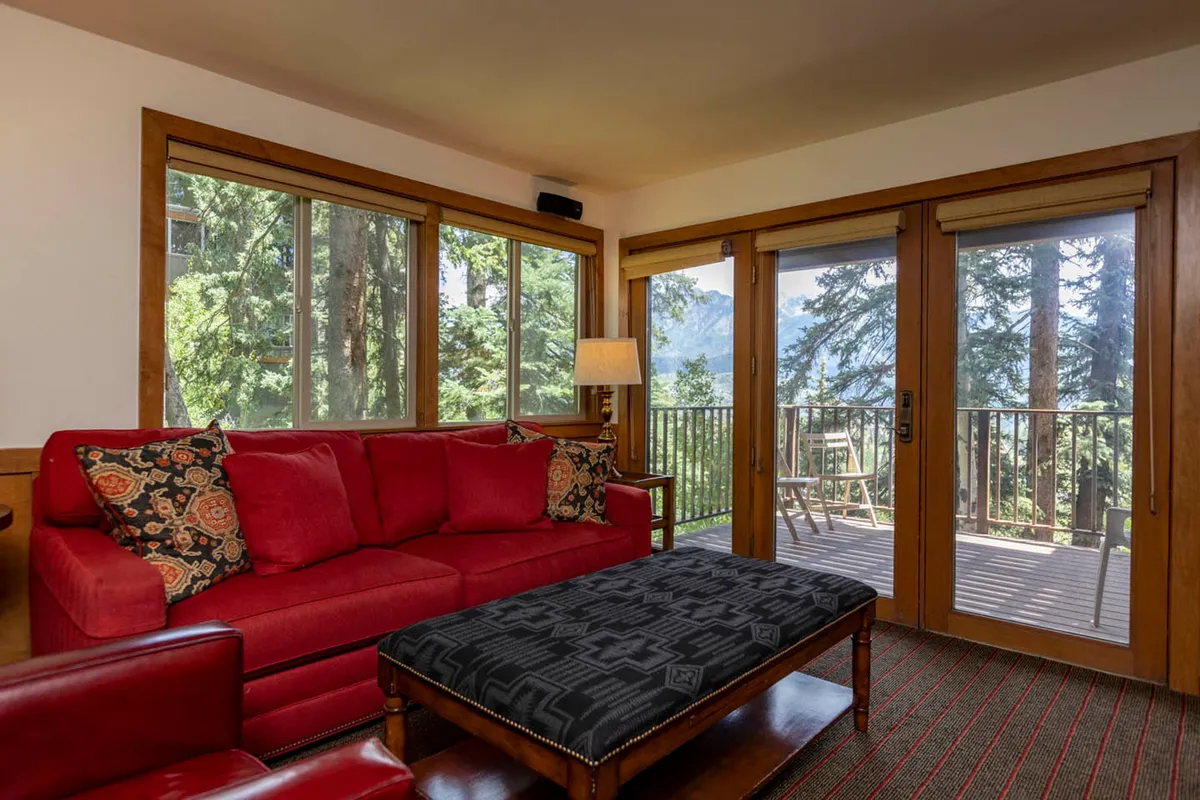 A bright, cheerful living space centered around a vivid red sofa with patterned pillows, large windows revealing impressive mountain and forest views, and a balcony equipped with outdoor chairs. The room feels both intimate and open, ideal for group relaxation, storytelling, and appreciating the surrounding nature together.