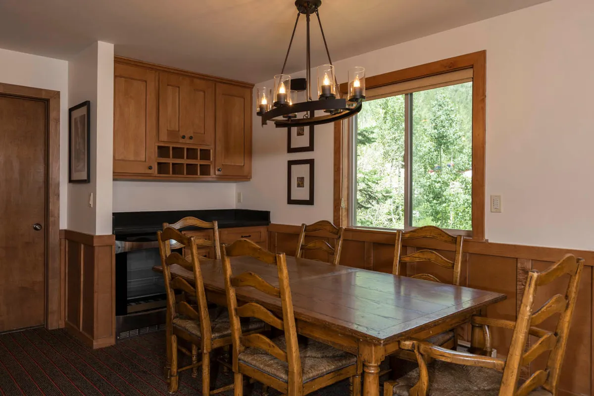 A rustic wooden dining table surrounded by chairs, set in front of a large window with a view of the forest. Warm light from a chandelier adds to the inviting ambiance, setting the scene for group meals, games, or planning the next day’s adventures.