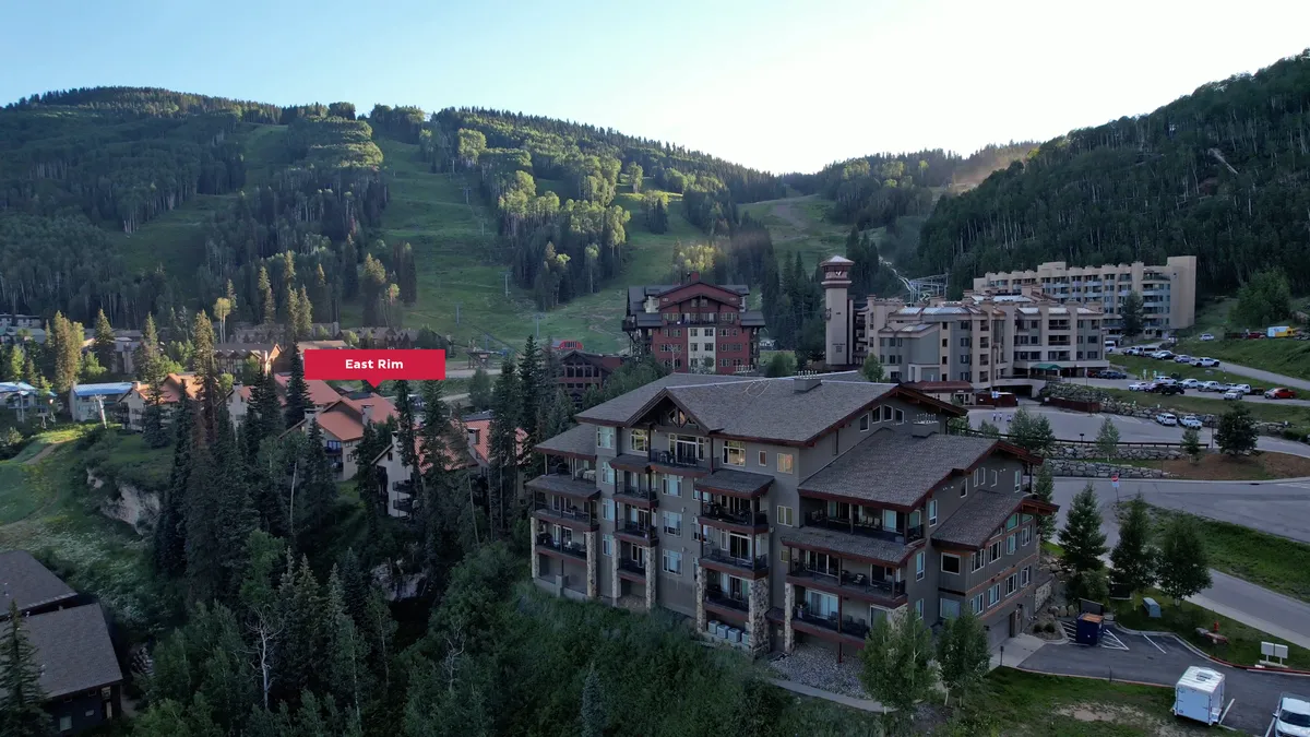 A multi-story condominium building with balconies stands prominently before ski trails marked on a hillside, surrounded by lush evergreen forest and adjacent resort buildings. The setting is tranquil, inviting, and clearly places the property right at the heart of the action.