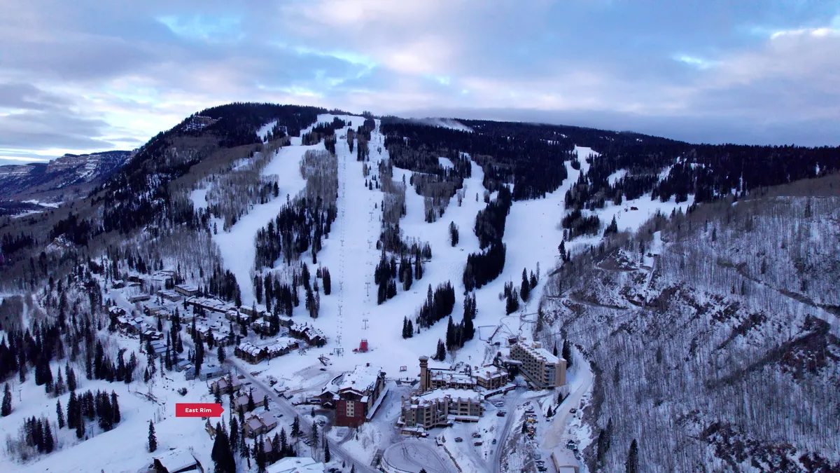 A breathtaking winter landscape with snow-covered ski slopes and a ski lift, surrounded by evergreen trees and resort buildings at the mountain base. The scene conveys easy access to outdoor adventures and the stunning mountain setting.