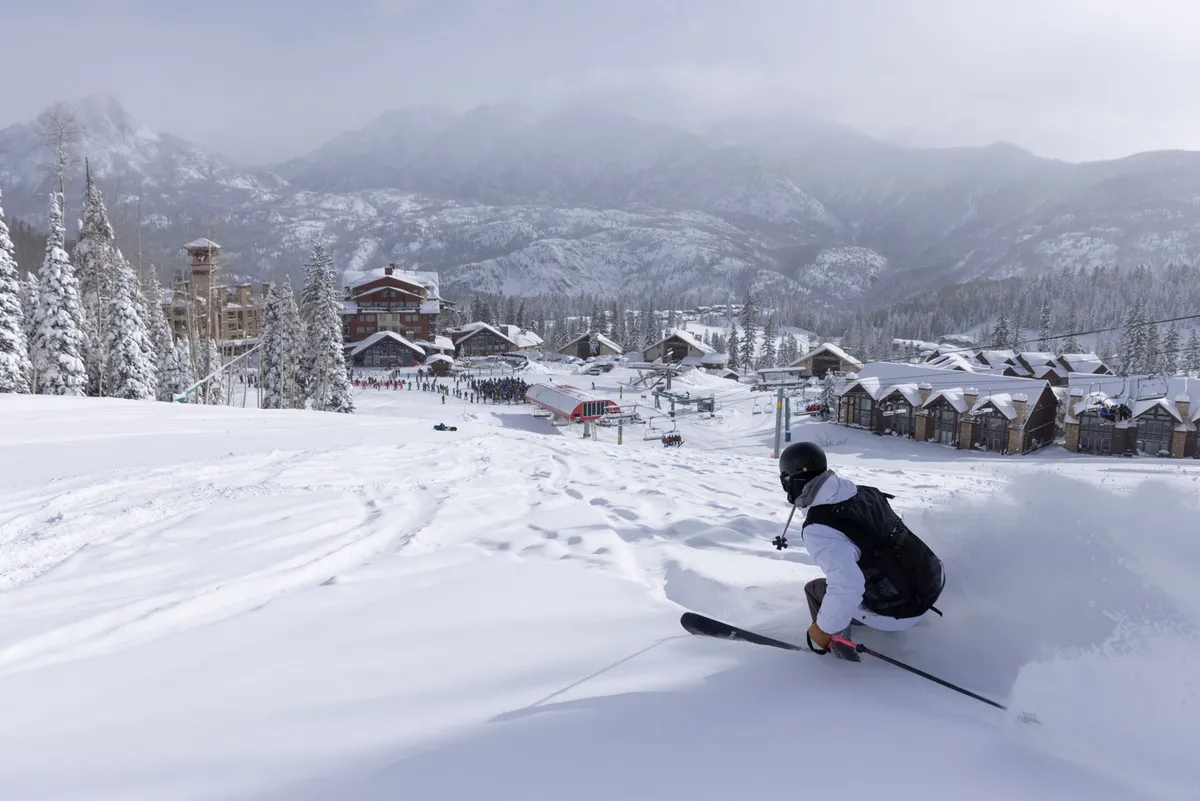 A skier carves through fresh snow on a pristine slope, with snow-blanketed ski runs, resort buildings nestled below, and distant misty mountains providing a dramatic winter backdrop. The action and scenery together evoke the exhilaration and beauty of mountain living.