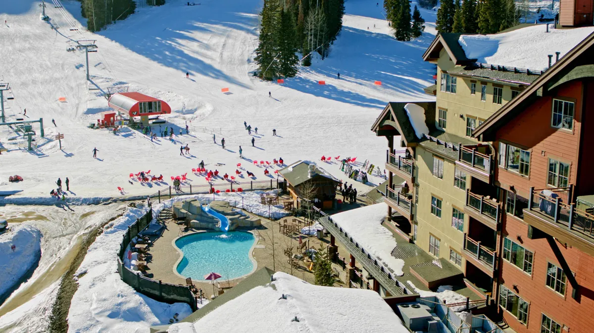 This image showcases skiers gliding down snow-covered slopes, an outdoor heated pool steaming amid the snow, a multi-story lodge building with balconies, and a ski lift with its vibrant red station. The scene is lively, capturing both the thrill of skiing and the relaxation of resort amenities against a backdrop of mountain beauty.