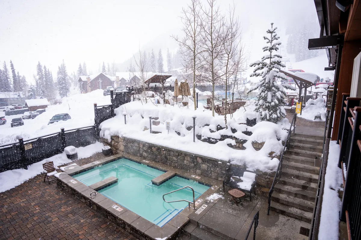 A serene winter scene featuring a heated outdoor pool surrounded by snow, with pine trees and mountain lodges in the background. Stone retaining walls and stairs are dusted with fresh snow, creating a peaceful, inviting ambiance. The snowfall adds a magical touch, and the scene hints at outdoor relaxation or play for families, even for non-skiers.