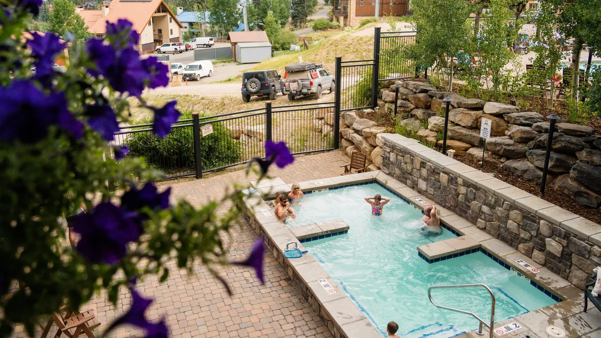 A lively outdoor pool scene with people of various ages enjoying the heated water. The area is landscaped with stone walls and vibrant purple flowers, enclosed by a black metal fence. The mood is joyful and communal, with children and adults alike socializing and relaxing together.
